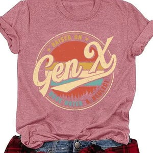 Puede incluir: Camiseta rosa con mangas enrolladas. Presenta un gr&aacute;fico circular con "Gen X" en una fuente retro, rodeado por "Raised on MTV & Neglect". Una camisa a cuadros roja y negra est&aacute; atada a la cintura. La camiseta se combina con unos vaqueros azules.