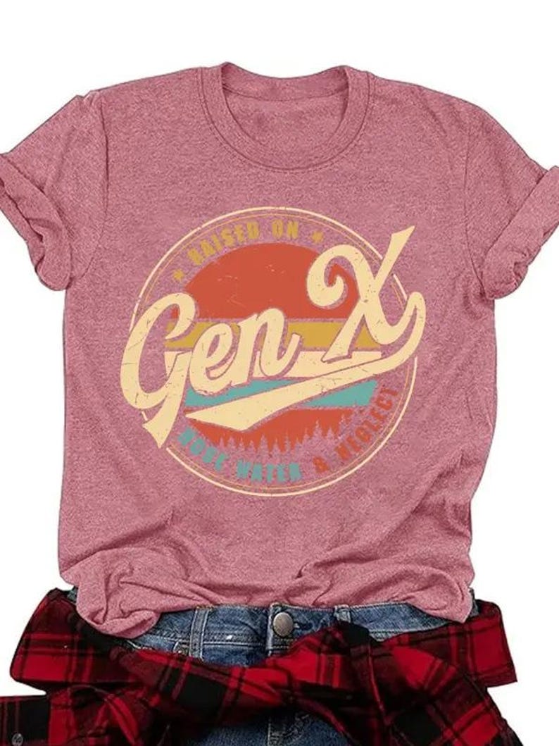 Puede incluir: Camiseta rosa con mangas enrolladas. Presenta un gr&aacute;fico circular con "Gen X" en una fuente retro, rodeado por "Raised on MTV & Neglect". Una camisa a cuadros roja y negra est&aacute; atada a la cintura. La camiseta se combina con unos vaqueros azules.