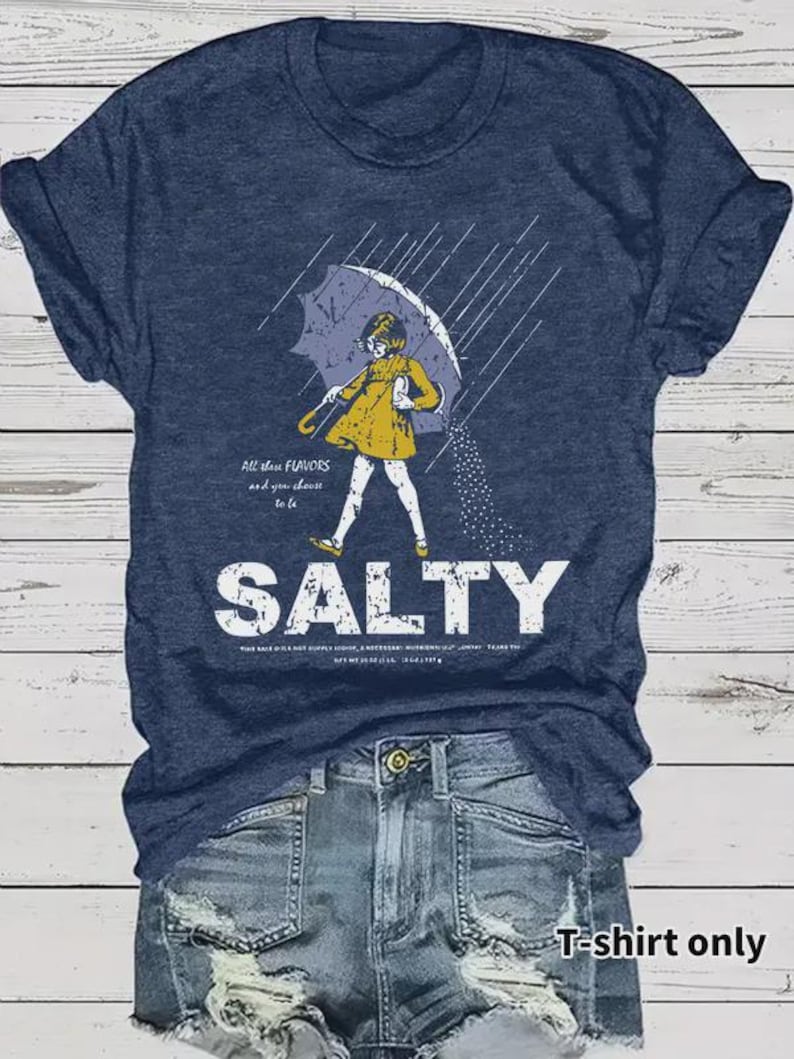 May include: Navy blue t-shirt with a graphic of a person under an umbrella, with the word "SALTY" in large white letters. The person is wearing a yellow coat. The text "All those flavors and you choose to be" is also on the shirt.