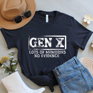 May include: A navy blue t-shirt with the text "GEN X LOTS OF MEMORIES NO EVIDENCE" in white. The shirt is paired with blue jeans, a brown hat, and sunglasses. A casual, everyday style.
