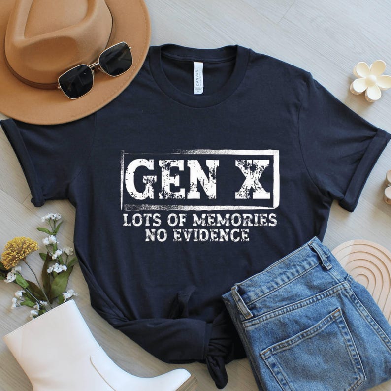 May include: A navy blue t-shirt with the text "GEN X LOTS OF MEMORIES NO EVIDENCE" in white. The shirt is paired with blue jeans, a brown hat, and sunglasses. A casual, everyday style.