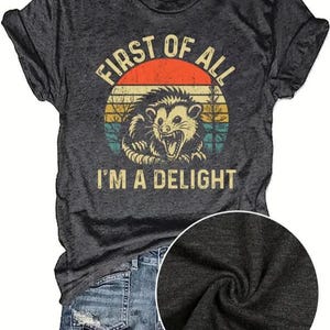 May include: Dark gray t-shirt with a distressed graphic of an opossum inside a sunset circle. The text reads "FIRST OF ALL I'M A DELIGHT". The shirt is paired with denim shorts.