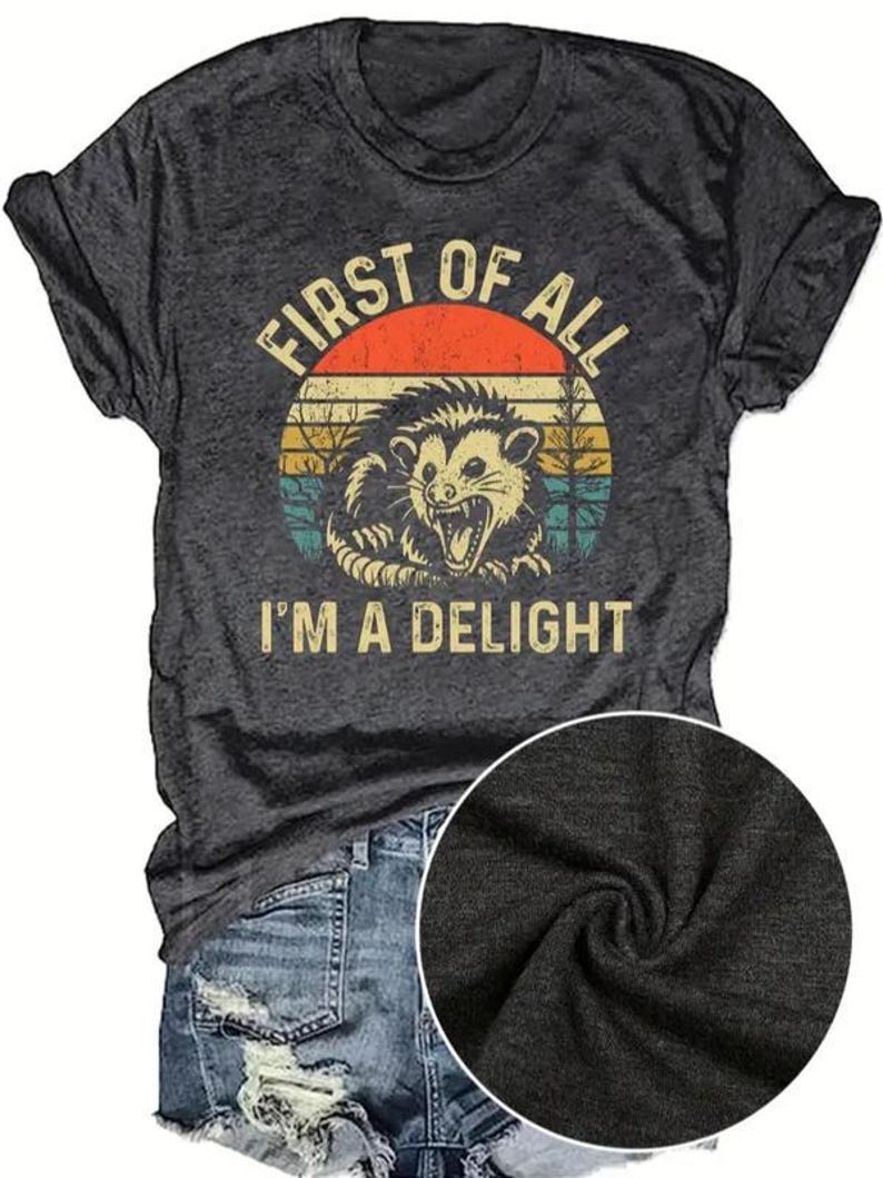 May include: Dark gray t-shirt with a distressed graphic of an opossum inside a sunset circle. The text reads "FIRST OF ALL I'M A DELIGHT". The shirt is paired with denim shorts.