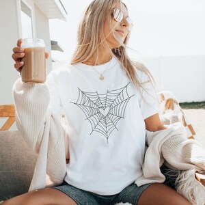 May include: White crew neck t-shirt with a black spiderweb design and a heart in the middle. The person in the image is wearing sunglasses and holding a glass of iced coffee. The shirt is a casual style.