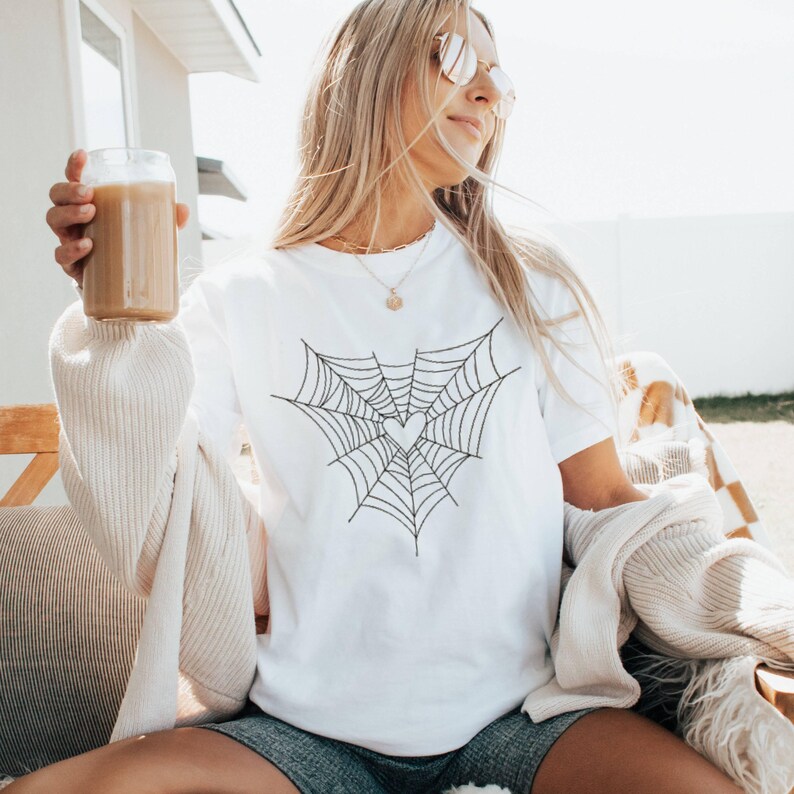 May include: White crew neck t-shirt with a black spiderweb design and a heart in the middle. The person in the image is wearing sunglasses and holding a glass of iced coffee. The shirt is a casual style.