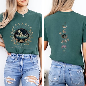 Puede incluir: Camiseta de color verde azulado con un diseño circular frontal con el texto "VELARIS" y una escena de montaña. La parte trasera de la camiseta presenta un diseño de fases lunares con mariposas y detalles florales. La camiseta es de material suave.