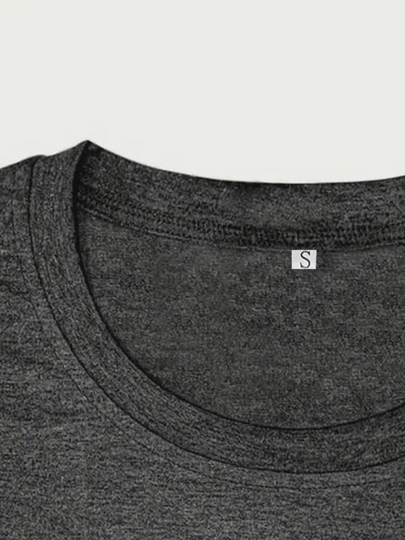 May include: Close-up of a dark gray t-shirt neckline. The shirt has a crew neck and a small white tag with the letter "S" on it. The fabric has a textured appearance.