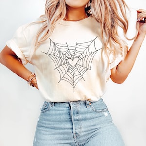 May include: A cream-coloured t-shirt with a black spiderweb design and a pink heart at the centre. The soft material t-shirt is suitable for casual wear. The design is centered on the front of the shirt.