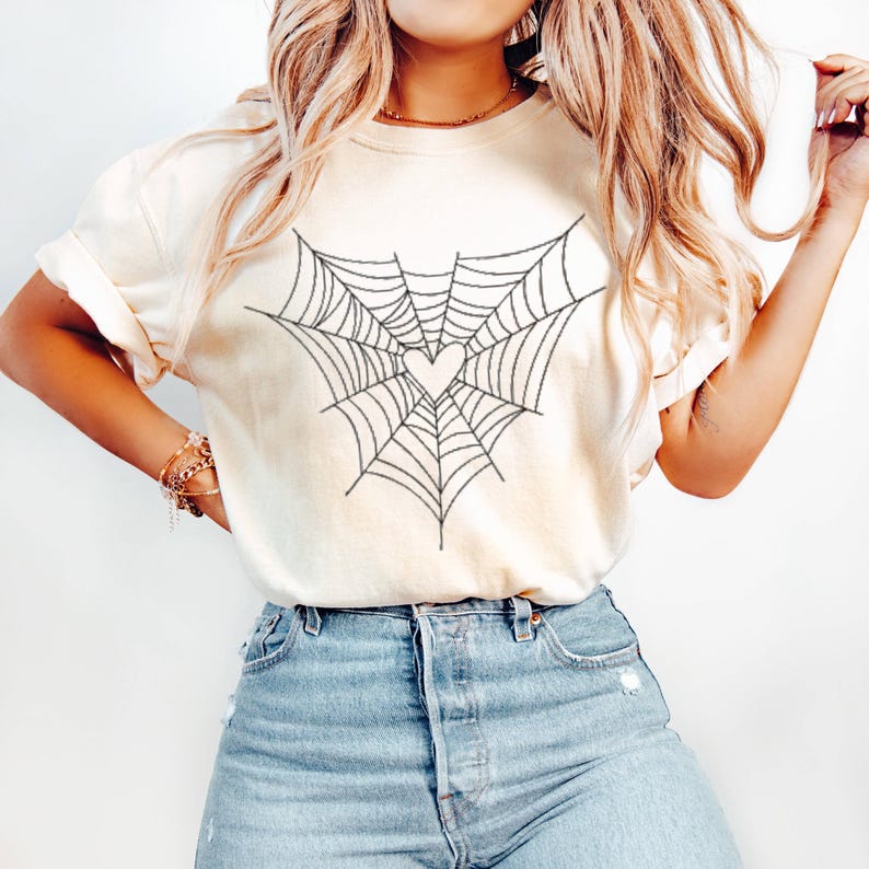 May include: A cream-coloured t-shirt with a black spiderweb design and a pink heart at the centre. The soft material t-shirt is suitable for casual wear. The design is centered on the front of the shirt.