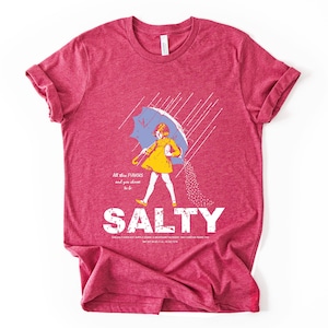 May include: A red t-shirt with a graphic of a person under a blue umbrella, with the word "SALTY" in large white letters. The person is wearing a yellow dress and is being sprinkled with salt. The text "All the flavors and you choose to be" is above the word "SALTY".