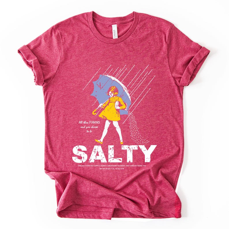 May include: A red t-shirt with a graphic of a person under a blue umbrella, with the word "SALTY" in large white letters. The person is wearing a yellow dress and is being sprinkled with salt. The text "All the flavors and you choose to be" is above the word "SALTY".