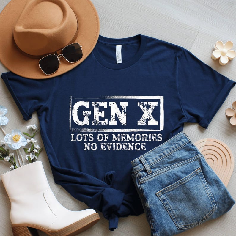 May include: A navy blue t-shirt with the text "GEN X LOTS OF MEMORIES NO EVIDENCE" in a white, distressed font. The shirt is paired with blue jeans, a brown hat, sunglasses, and white boots.