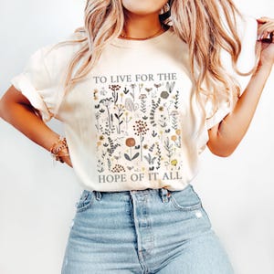 May include: Cream-colored t-shirt with a floral design and the text "TO LIVE FOR THE HOPE OF IT ALL". The graphic features various botanical illustrations in muted tones, including flowers and leaves. The shirt is a casual, comfortable style.