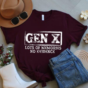 May include: A maroon t-shirt with the text "GEN X LOTS OF MEMORIES NO EVIDENCE" in a white rectangular box. The shirt is paired with blue jeans, a brown hat, sunglasses, and white boots. The shirt is made of a soft material.