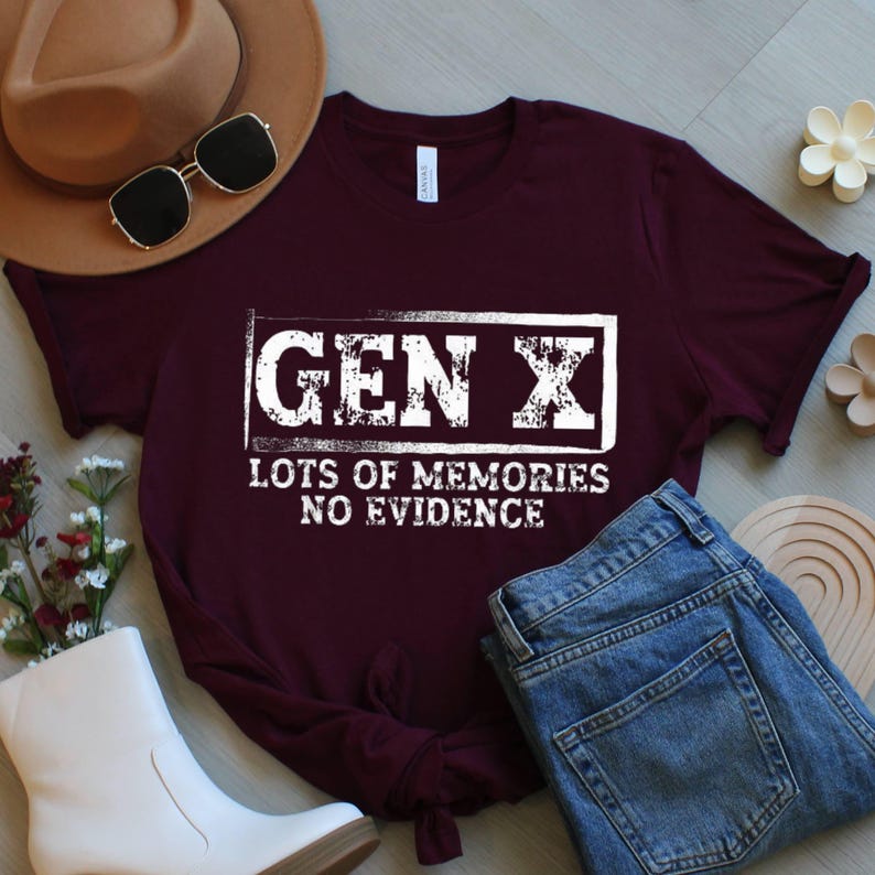 May include: A maroon t-shirt with the text "GEN X LOTS OF MEMORIES NO EVIDENCE" in a white rectangular box. The shirt is paired with blue jeans, a brown hat, sunglasses, and white boots. The shirt is made of a soft material.