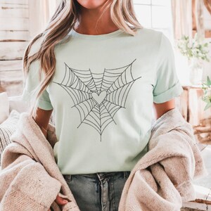 May include: A pale green t-shirt with a black spiderweb design incorporating a heart shape. The short-sleeved shirt is made of a soft material, suitable for casual wear. The shirt is a solid pale green colour.