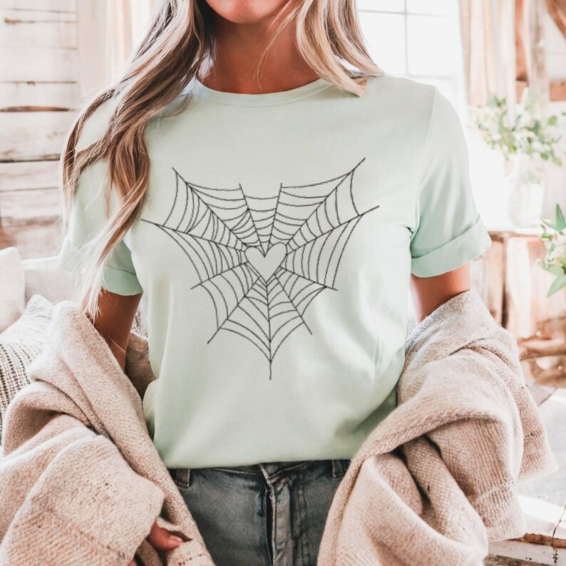 May include: A pale green t-shirt with a black spiderweb design incorporating a heart shape. The short-sleeved shirt is made of a soft material, suitable for casual wear. The shirt is a solid pale green colour.