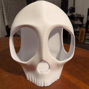 Puede incluir: Una máscara de calavera impresa en 3D de color blanco con grandes cuencas oculares huecas y redondeadas y una pequeña abertura nasal triangular. La superficie muestra sutiles líneas de capa horizontales características de la fabricación aditiva.