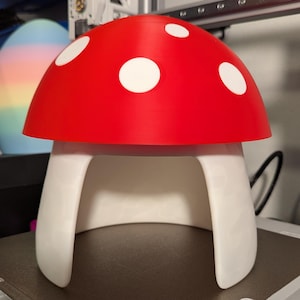 Op de afbeelding: Een paddestoelvormig object met een felrode koepelvormige hoed versierd met verschillende witte cirkelvormige stippen. De basis is wit en gebogen, wat een open boogstructuur vormt. Het item heeft een gladde, matte afwerking en een symmetrisch ontwerp.