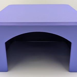 Puede incluir: Un soporte de exhibición sólido de color violeta claro con un acabado liso y mate. La estructura presenta una superficie superior plana y cuadrada con esquinas redondeadas y un corte arqueado ancho centrado en el panel frontal.