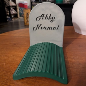 Può includere: Un supporto grigio a forma di lapide con il testo "Abby Normal" in rilievo sulla parte anteriore. La base è una piattaforma scanalata verde scuro che si estende in avanti, progettata per sostenere un oggetto in verticale contro il pannello.