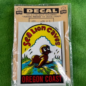 May include: A vintage decal featuring a cartoon seal with the text "Sea Lion Caves Oregon Coast." The decal is in a plastic sleeve with a brown backing that reads "DECAL" and "IMPKO."