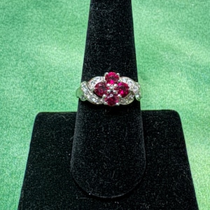 May include: A silver ring with a cluster of red gemstones in a floral design, surrounded by smaller clear stones. The ring is displayed on a black ring holder against a green background.
