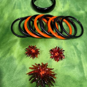 May include: A collection of jewelry and accessories, including a black bangle bracelet, a set of black and orange bangle bracelets, and three red and orange flower-shaped brooches and earrings. The items are displayed on a green surface.