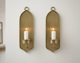 May include: Two brass wall sconces with a candle holder and a lit white candle. The sconces have an arched top and a decorative bottom. The sconces are mounted on a white wall.