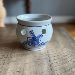 May include: White ceramic pot with blue windmill design. The pot has two circular holes and a blue trim around the top and base. The windmill design is on the side of the pot.