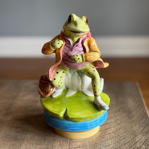 May include: A green frog figurine wearing a pink and yellow jacket, holding a basket, and standing on a green lily pad. The frog is a musical figurine and has a blue base.