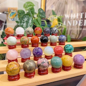 May include: A collection of 24 polished gemstone spheres displayed on wooden shelves. The spheres are various colours, including red, green, purple, blue, and brown. Each sphere is placed on a small wooden stand.