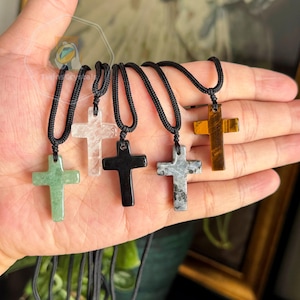 May include: A collection of five gemstone cross pendants on black cords. The crosses are in various colors, including green, clear, black, gray, and amber. The pendants are displayed against a neutral background.