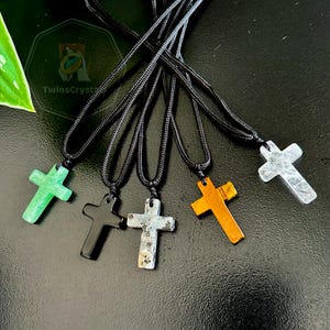 Peut inclure: Une collection de cinq pendentifs croix en pierres pr&eacute;cieuses sur des cordons noirs. Les croix sont de diff&eacute;rentes couleurs, dont le vert, le noir, le gris, l'orange et le transparent. Les pendentifs sont pr&eacute;sent&eacute;s sur un fond sombre.