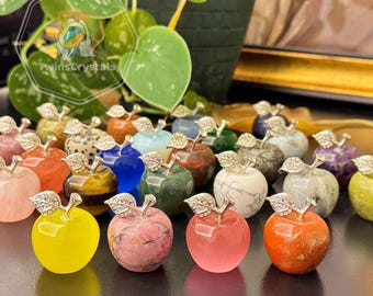 May include: A collection of gemstone apple figurines in various colors, including yellow, pink, blue, and green. Each apple has a silver-colored stem and leaf design. The background includes a plant with green leaves.