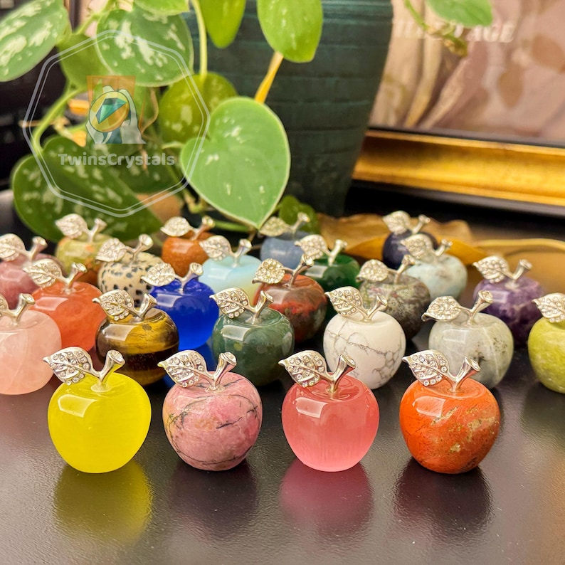 May include: A collection of gemstone apple figurines in various colors, including yellow, pink, blue, and green. Each apple has a silver-colored stem and leaf design. The background includes a plant with green leaves.