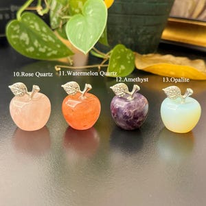 May include: Four gemstone apple sculptures. The apples are made of rose quartz, watermelon quartz, amethyst, and opalite. Each apple has a silver-colored stem and leaf design. The text on the image reads: "10. Rose Quartz", "11. Watermelon Quartz", "12. Amethyst", and "13. Opalite".