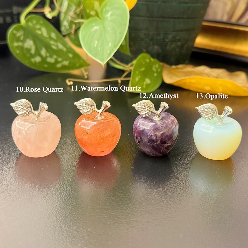 May include: Four gemstone apple sculptures. The apples are made of rose quartz, watermelon quartz, amethyst, and opalite. Each apple has a silver-colored stem and leaf design. The text on the image reads: "10. Rose Quartz", "11. Watermelon Quartz", "12. Amethyst", and "13. Opalite".
