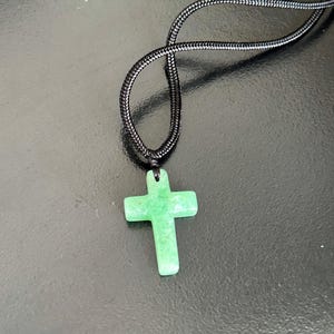 Peut inclure: Pendentif croix en pierre verte sur un collier en cordon noir. La croix est de couleur vert clair et a une surface lisse. Le cordon du collier est en mat&eacute;riau noir tiss&eacute; serr&eacute;.