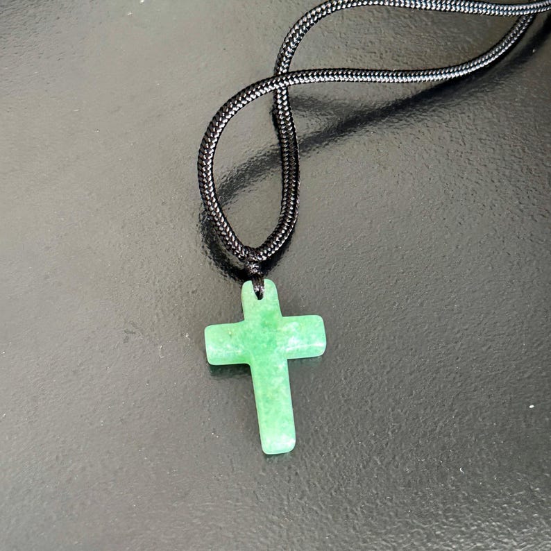 Peut inclure: Pendentif croix en pierre verte sur un collier en cordon noir. La croix est de couleur vert clair et a une surface lisse. Le cordon du collier est en mat&eacute;riau noir tiss&eacute; serr&eacute;.