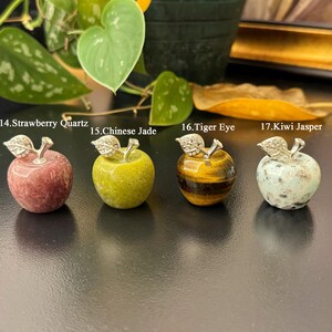 May include: Four decorative apple figurines made of different stones: Strawberry Quartz, Chinese Jade, Tiger Eye, and Kiwi Jasper. Each apple has a silver-colored leaf and stem. The apples are displayed on a black surface.