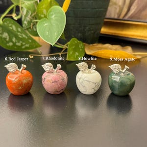 May include: Four gemstone apple sculptures. The apples are made of red jasper, rhodonite, howlite, and moss agate. Each apple has a silver-colored metal stem and leaf design. The apples are displayed on a dark surface.