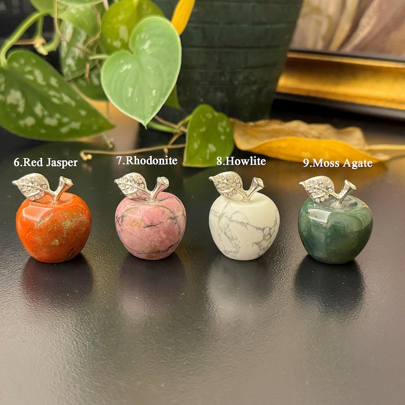 May include: Four gemstone apple sculptures. The apples are made of red jasper, rhodonite, howlite, and moss agate. Each apple has a silver-colored metal stem and leaf design. The apples are displayed on a dark surface.