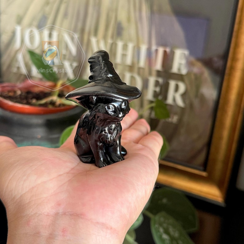 May include: A glossy black cat figurine wearing a witch's hat. The cat is sitting upright with its front paws together. The figurine is held in a person's hand, with a blurred background.