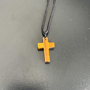 Peut inclure: Un pendentif en forme de croix en bois avec un motif &agrave; rayures marron et or est suspendu &agrave; un cordon noir. La croix est de conception simple, et le cordon semble &ecirc;tre fait de mat&eacute;riau tiss&eacute;. Le pendentif mesure environ 3,8 cm de long.