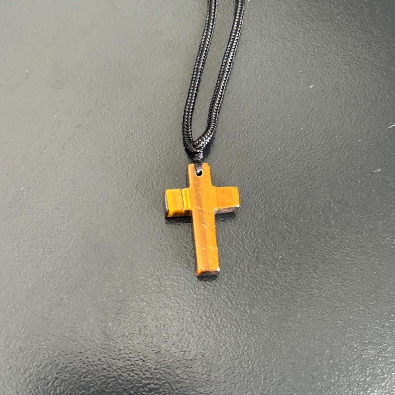 Peut inclure: Un pendentif en forme de croix en bois avec un motif &agrave; rayures marron et or est suspendu &agrave; un cordon noir. La croix est de conception simple, et le cordon semble &ecirc;tre fait de mat&eacute;riau tiss&eacute;. Le pendentif mesure environ 3,8 cm de long.
