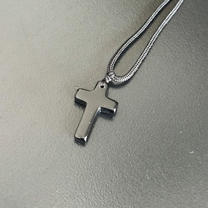 Peut inclure: Un collier avec un pendentif en forme de croix noire. La croix est noire, polie et suspendue &agrave; un cordon noir assorti. Le collier est pr&eacute;sent&eacute; sur une surface sombre et r&eacute;fl&eacute;chissante.