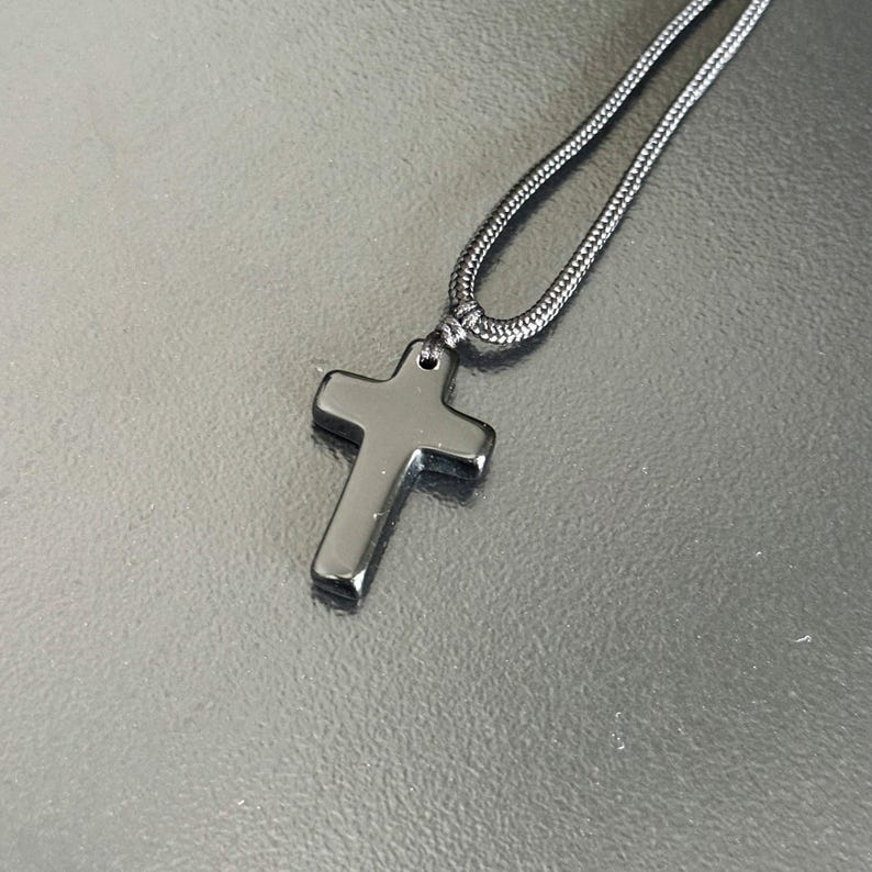 Peut inclure: Un collier avec un pendentif en forme de croix noire. La croix est noire, polie et suspendue &agrave; un cordon noir assorti. Le collier est pr&eacute;sent&eacute; sur une surface sombre et r&eacute;fl&eacute;chissante.