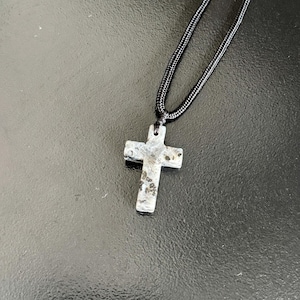 Peut inclure: Un collier avec un pendentif en forme de croix en pierre et un cordon noir. La croix est grise avec des mouchetures noires et marron. Un accessoire religieux.
