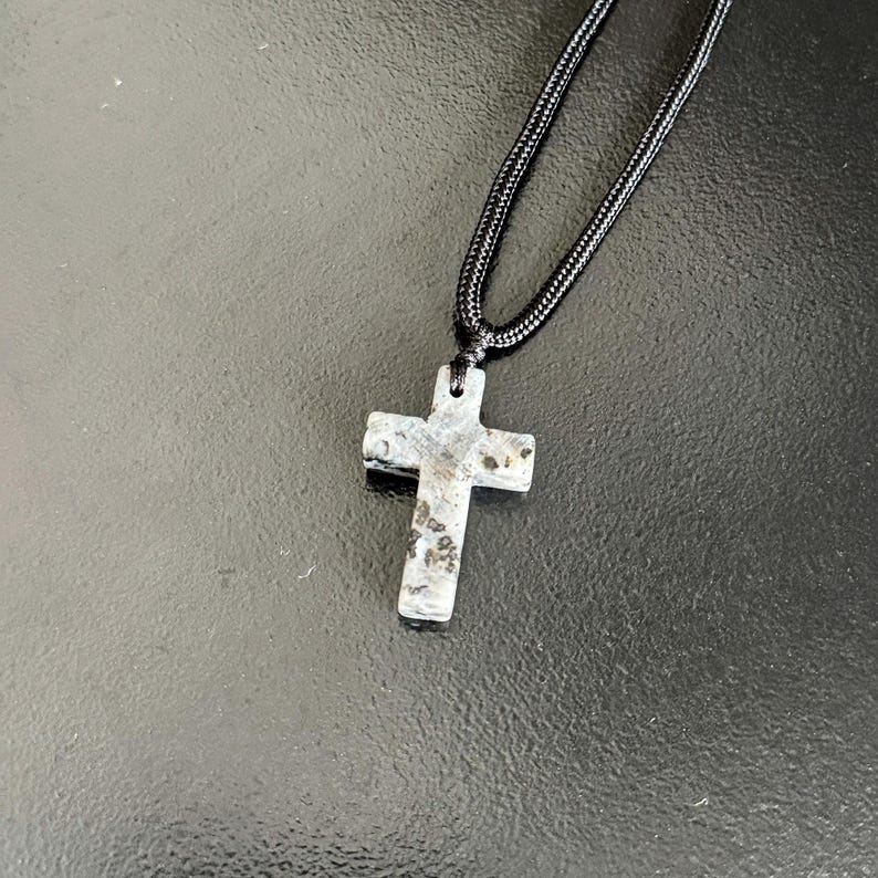 Peut inclure: Un collier avec un pendentif en forme de croix en pierre et un cordon noir. La croix est grise avec des mouchetures noires et marron. Un accessoire religieux.
